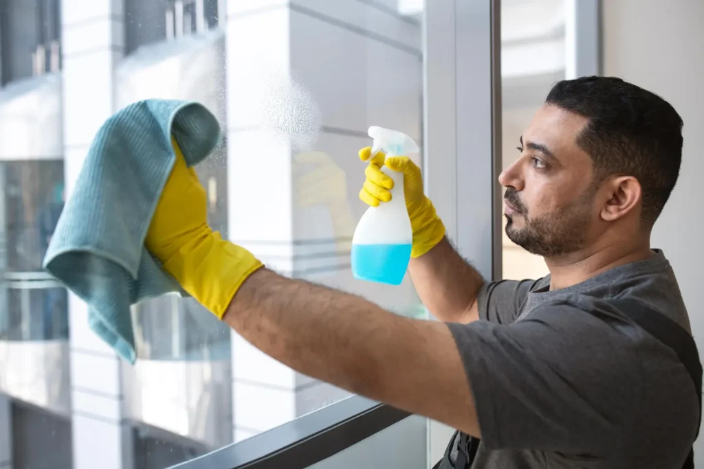 Man wearing yellow gloves spraying and wiping a large window with blue cleaner