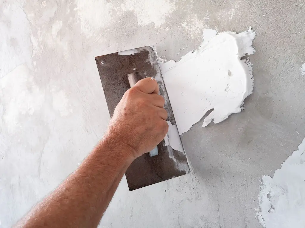 Hand using a wide trowel to spread white wall putty on a plastered wall.