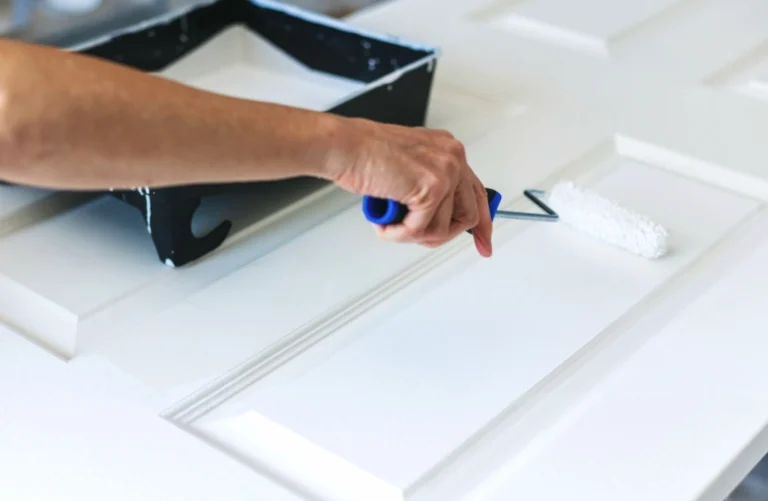 Hand using a paint roller to apply white paint to a door.Cracked grout line between wall and floor tiles.