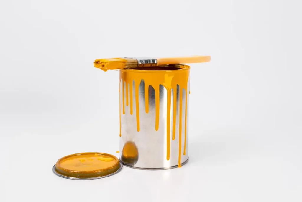 Open paint can with yellow paint dripping down the sides and a brush resting on top.