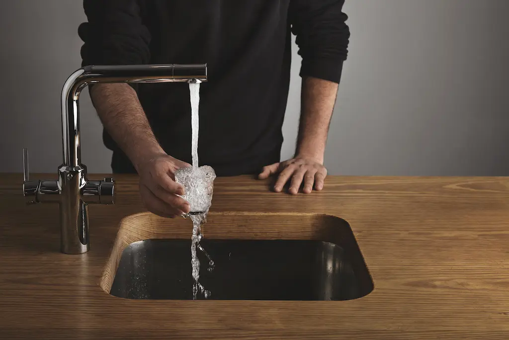 “Person filling a glass with running water at a modern chrome kitchen faucet over a wood countertop