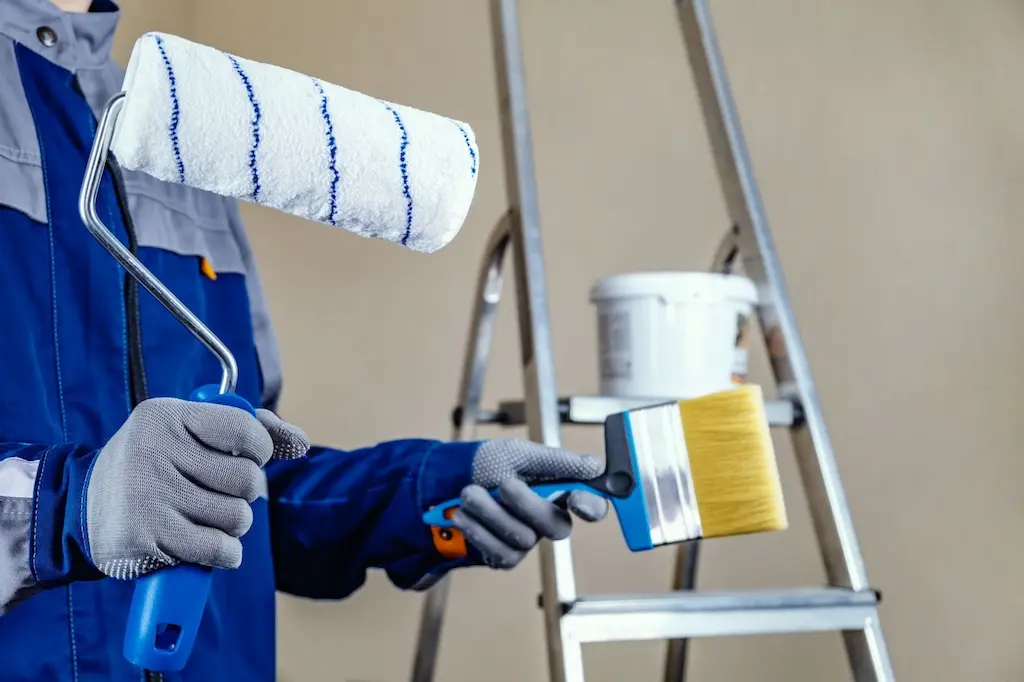 Painter in workwear holding a paint roller and wide brush with a ladder and paint bucket in the background.