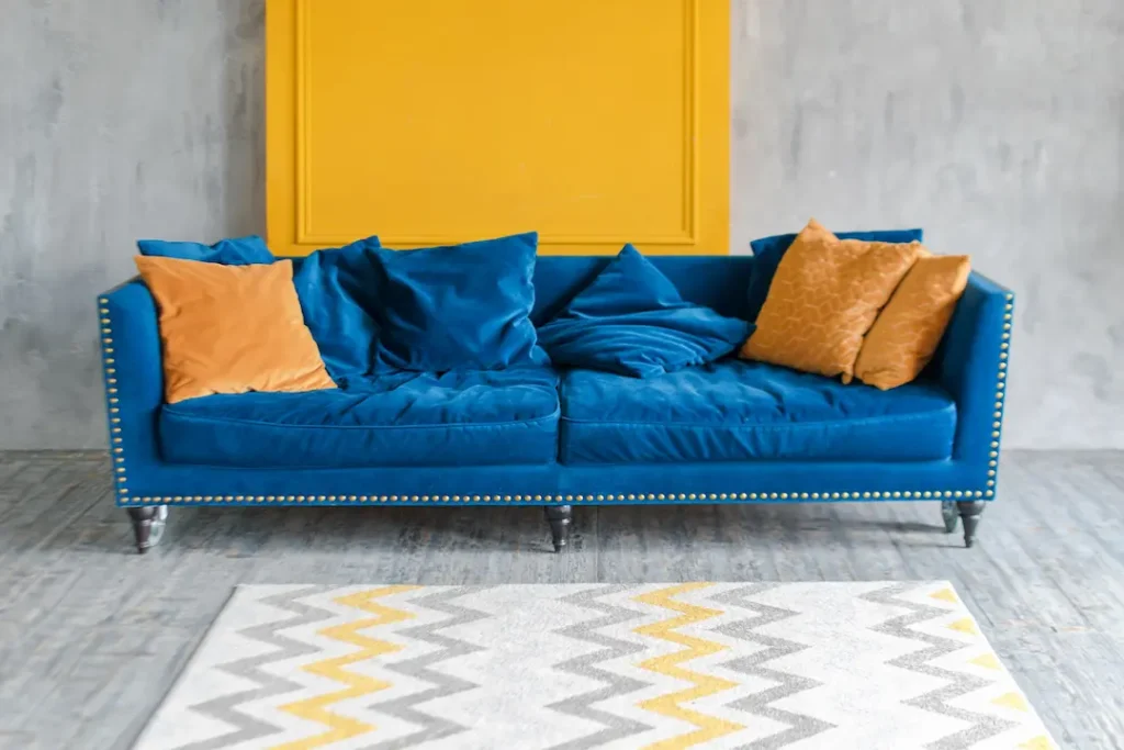 Bright blue velvet sofa with mustard-yellow throw pillows against a gray wall and yellow panel, with a chevron rug on the floor.